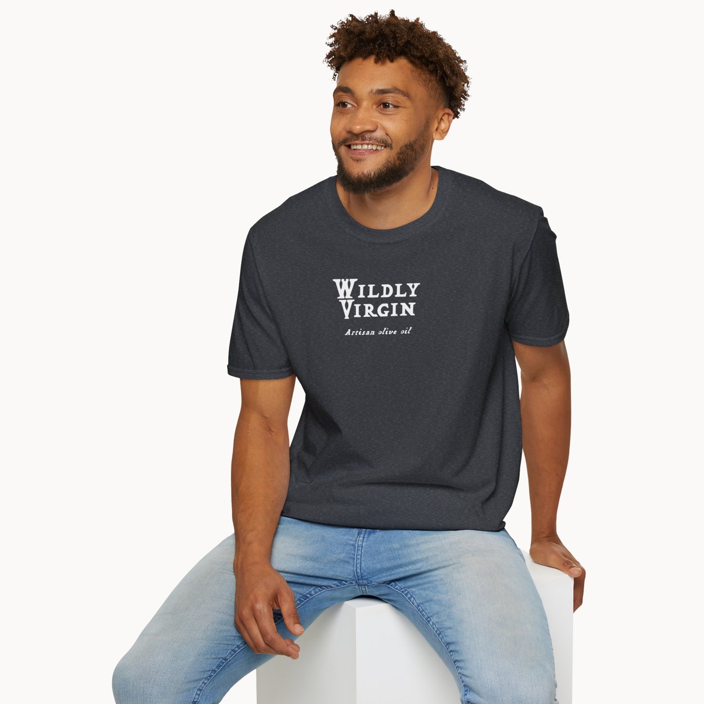 Man wearing a dark t-shirt with the text 'WILDLY VIRGIN' and a relaxed fit, sitting on a white cube.