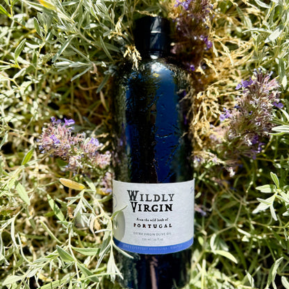 Bottle of Wildly Virgin extra virgin olive oil from Portugal, surrounded by herbs and flowers.