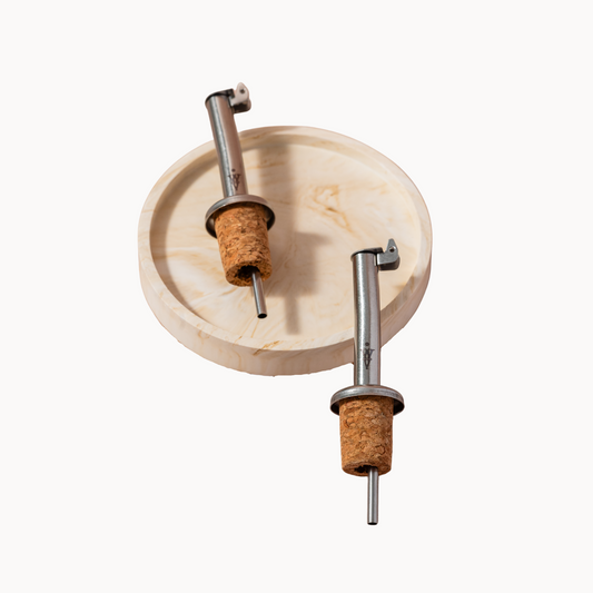 Two stainless steel olive oil pour spouts with cork bases on a round wooden tray, designed for perfect pouring.