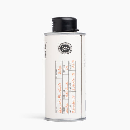 A vintage-style metal canister with a black screw cap, featuring a cream-colored label with handwritten details and a circular logo, set against a plain white background.