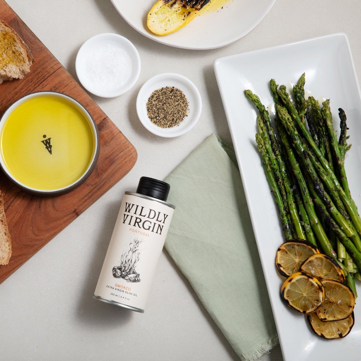 A styled flat-lay featuring a can of Wildly Virgin olive oil surrounded by food props including sliced rustic bread on a wooden board, grilled asparagus on a white platter, a bowl of yellow dip, and small bowls of salt and pepper.