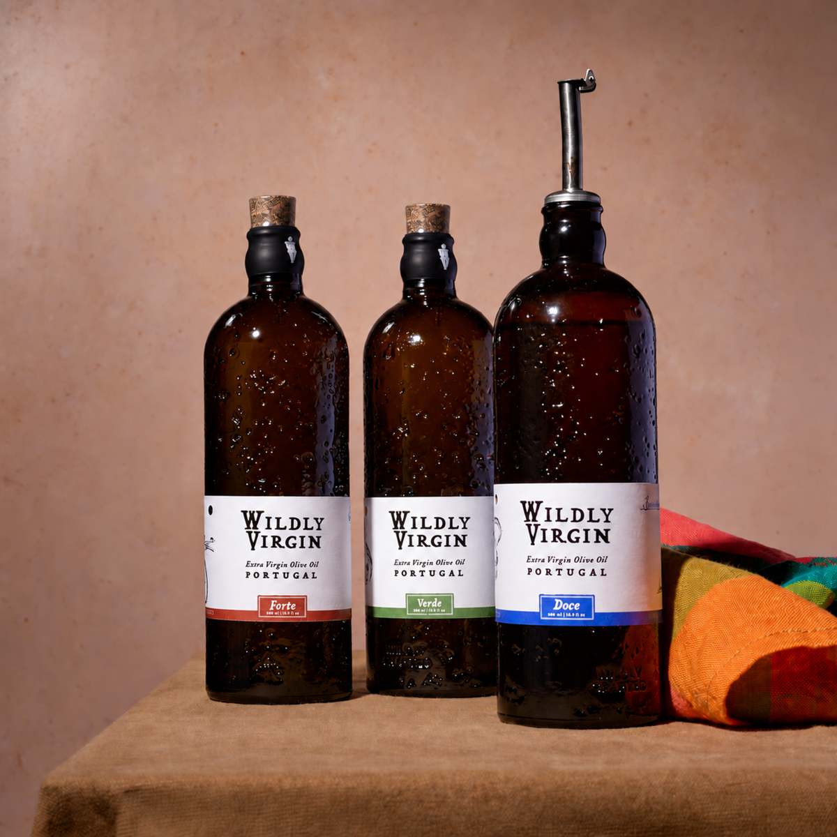 Three dark amber glass bottles of Wildly Virgin Portuguese olive oil with cork stoppers and a metal pour spout, arranged on a draped table against a textured beige wall.