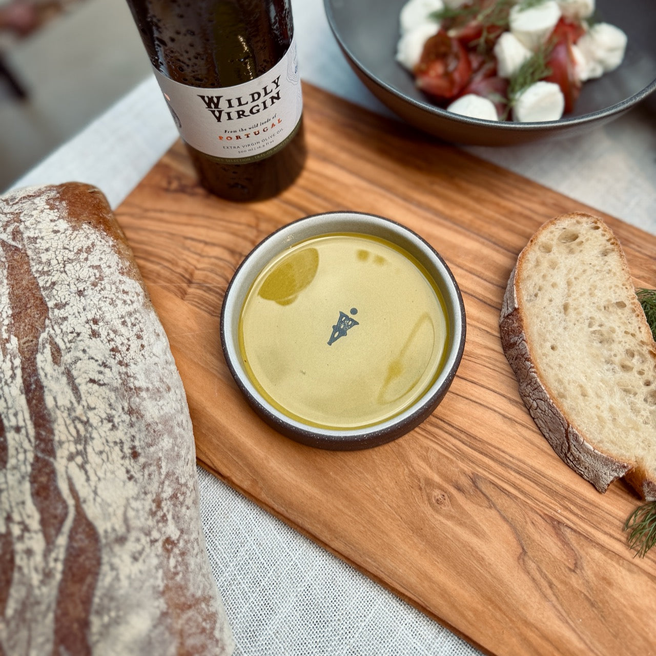 The Olive Oil Dish elegantly showcasing olive oil, accompanied by artisan bread and a bottle of wine on a wooden board.
