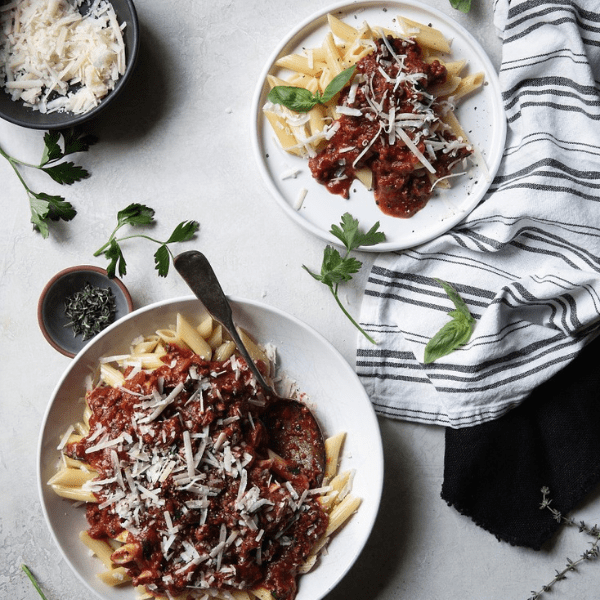 Two plates of pasta with marinara sauce and cheese, garnished with herbs on a textured surface.